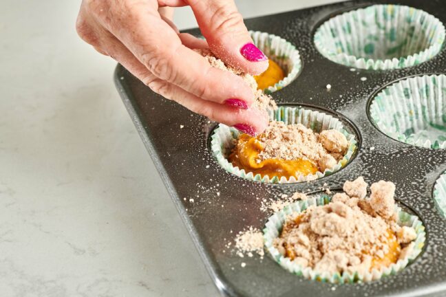 Woman sprinkling streusel topping onto muffin batter.