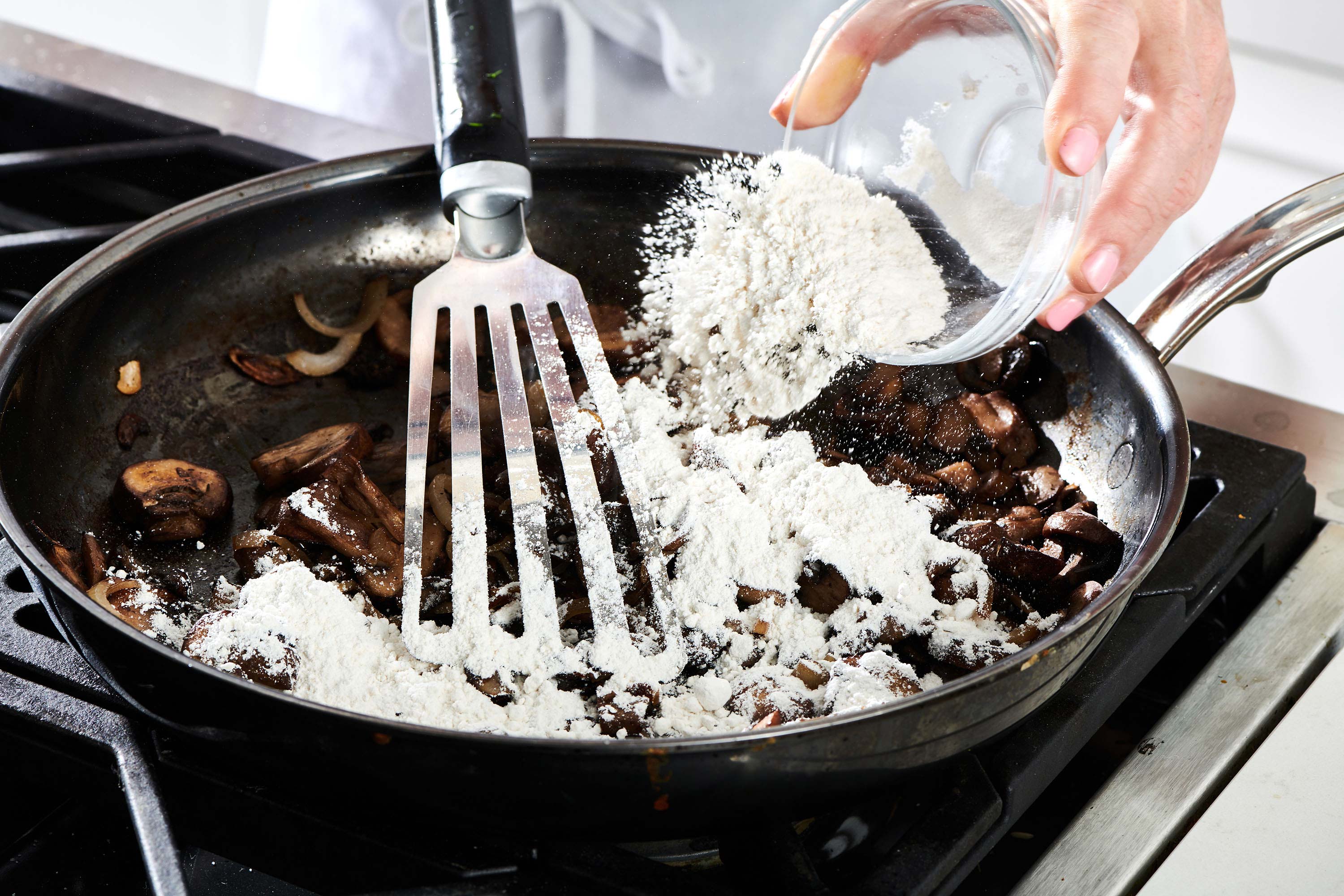 Woman sprinkling flour into a skillet with mushrooms and shallots.