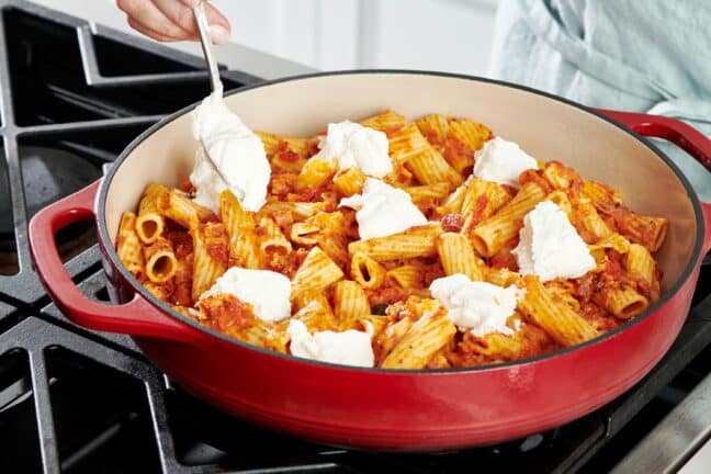 Spoon adding dollops of ricotta to a pan of noodles and sauce.