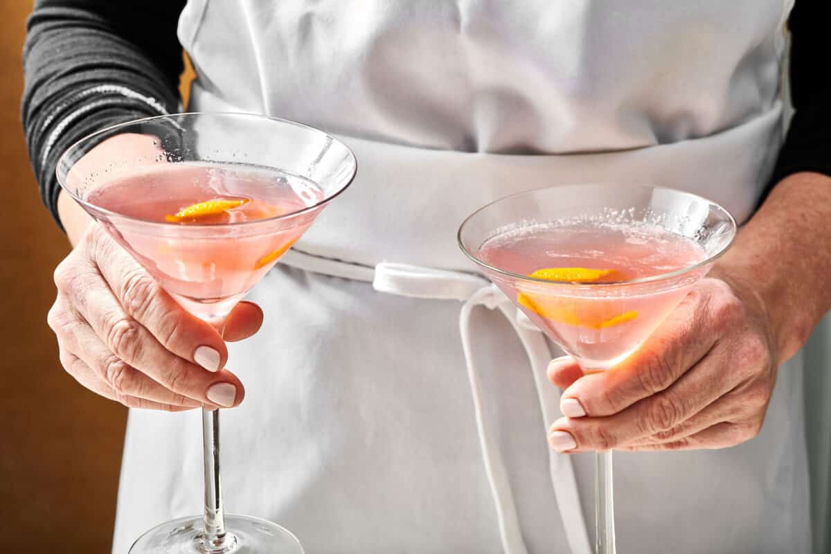 Woman holding two Cosmo Cocktails in long-stemmed glasses.