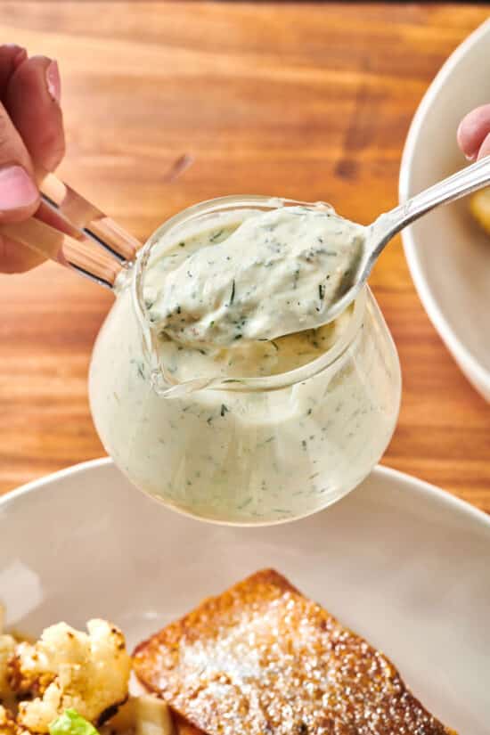 Spooning freshly made dill sauce from a jar over plate with poached salmon.