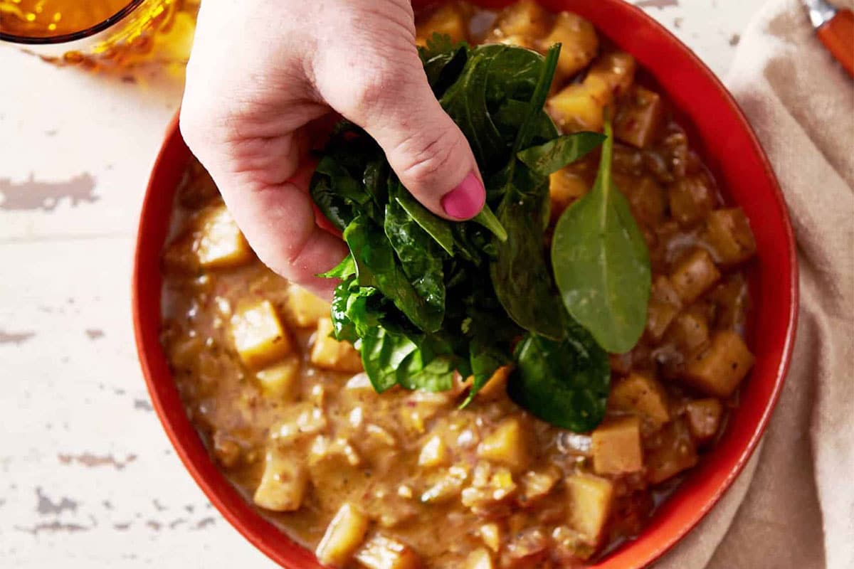 Woman adding spinach to a bowl of Potato Curry.
