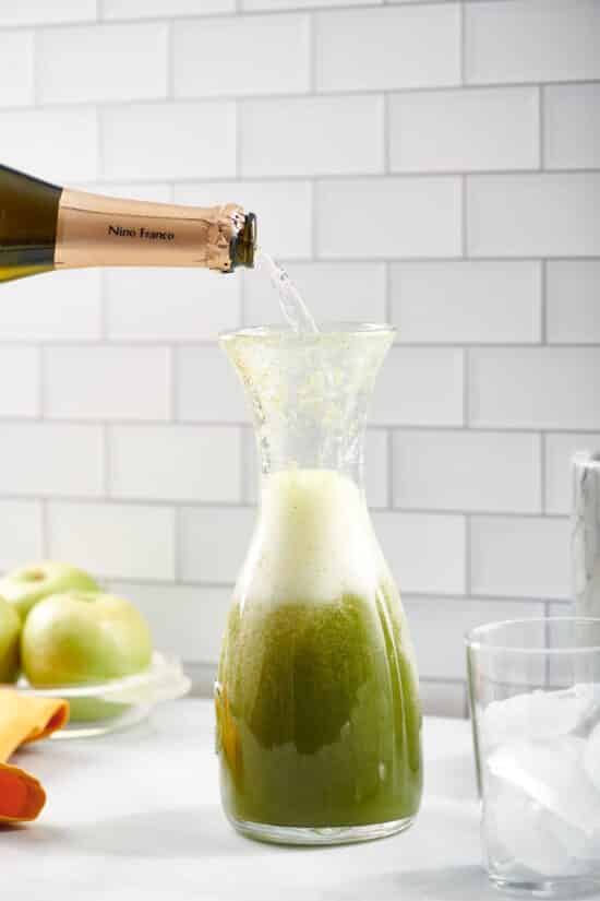 Bottle pouring Prosecco into a glass pitcher of green liquid.