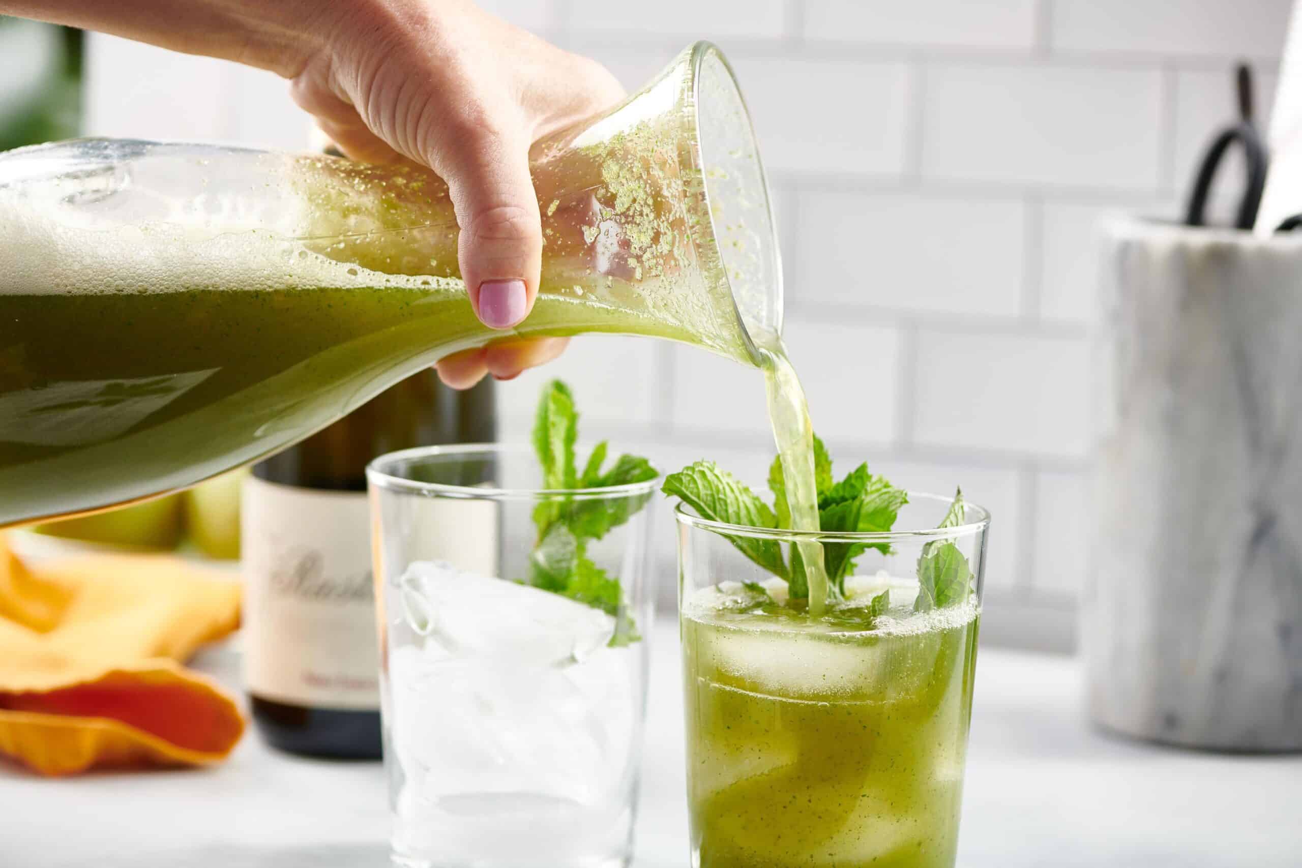 Glass pitcher pouring green Vodka Prosecco cocktails into small glasses of ice.