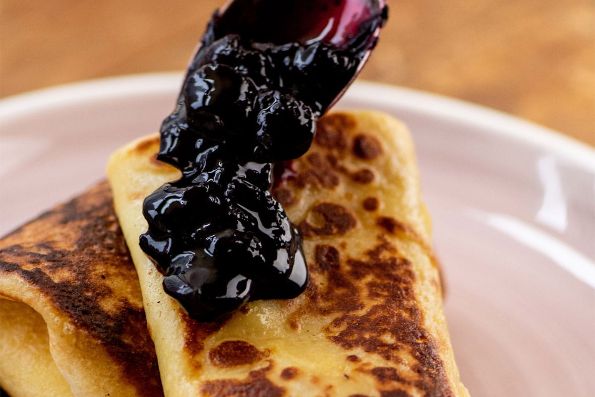 Spoon drizzling blueberry sauce over fresh blintze crepe.