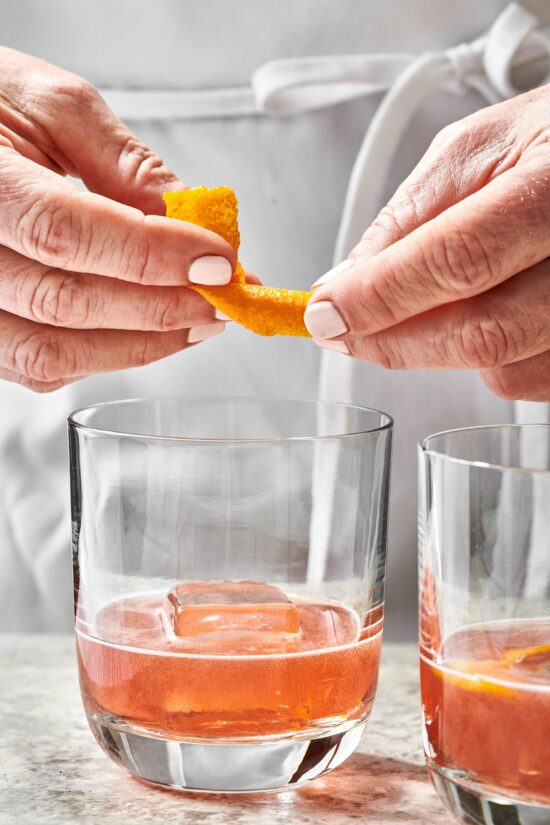 Woman placing orange peel into a Boulevardier Cocktail.
