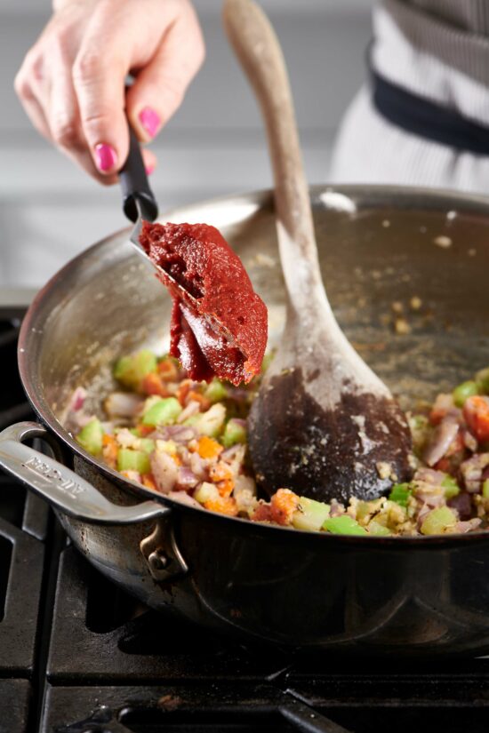 Spatula adding tomato paste to a skillet of diced vegetables.