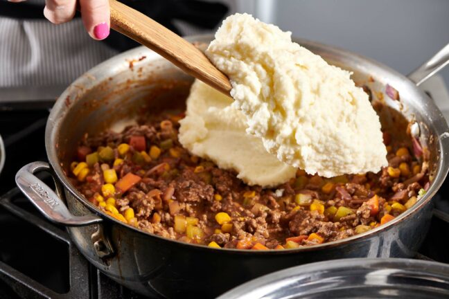 Woman adding mashed potatoes on top of a pan of vegetable and meat mixture.
