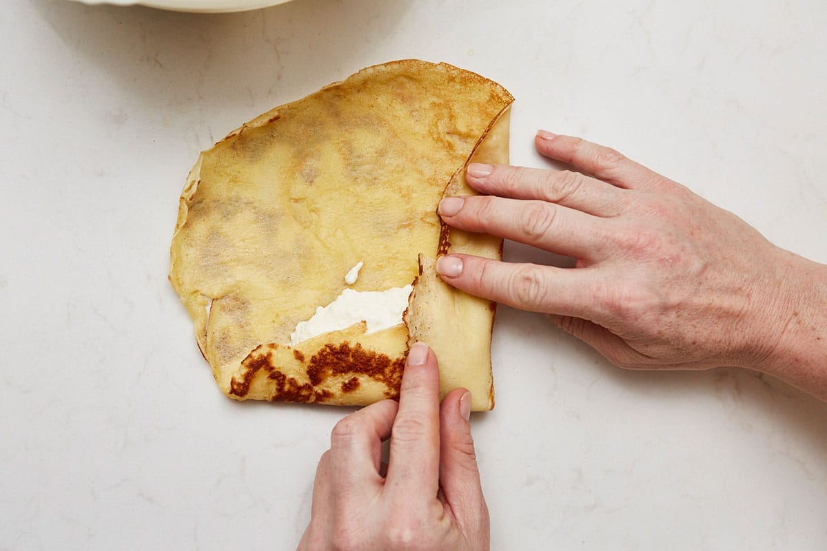 Woman folding the bottom and left sides on a crepe over the cheese filling.