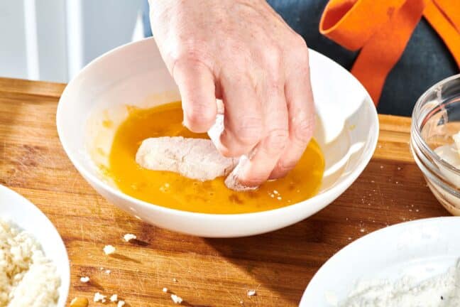 Woman placing a piece of floured fish into an egg mixture.