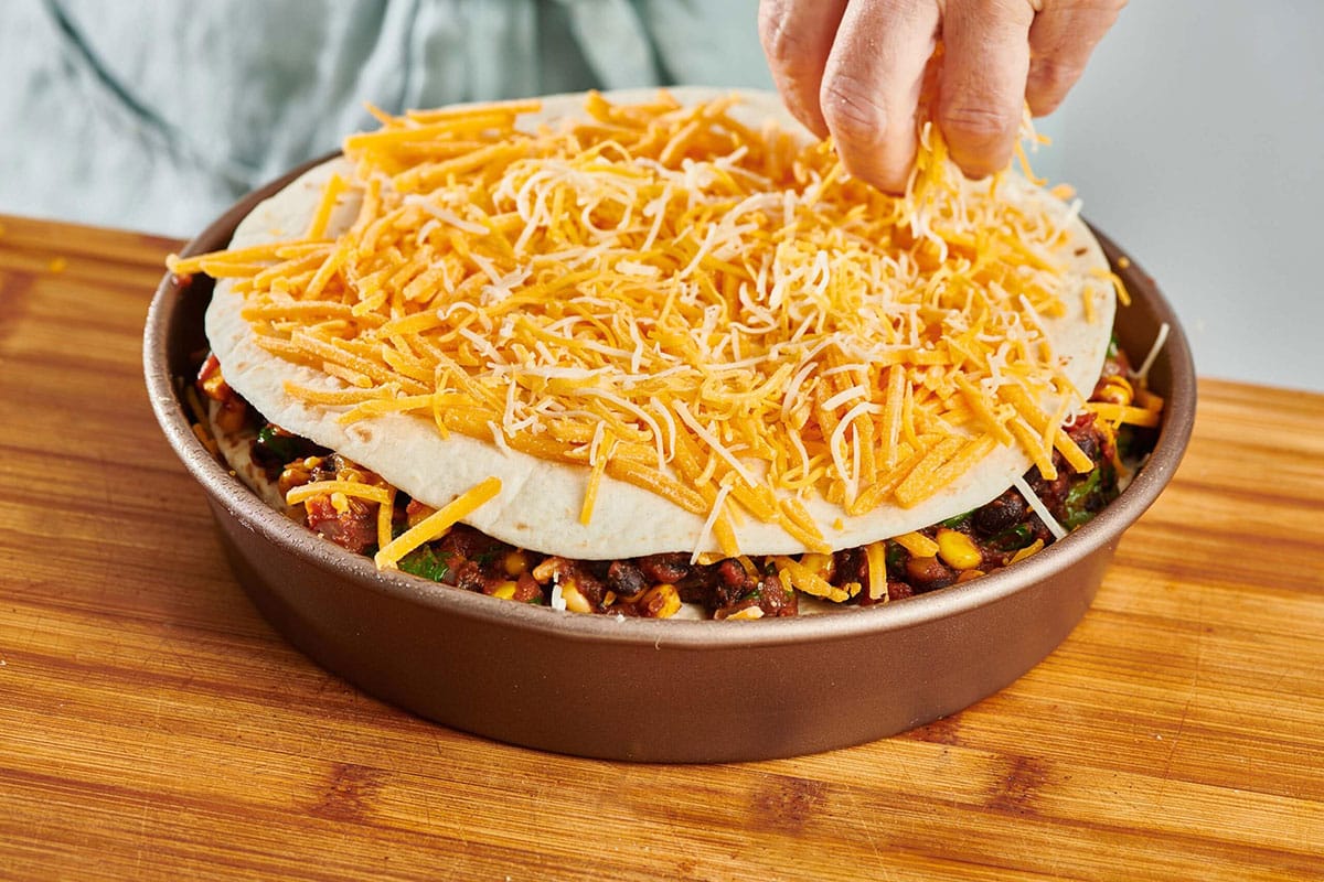 Layering cheese on tortilla and other Mexican lasagna ingredients in pan.