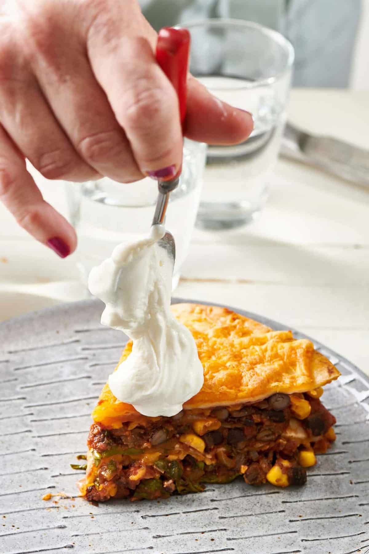 Spoon putting a dollop of sour cream on a slice of Vegetarian Mexican Lasagna.