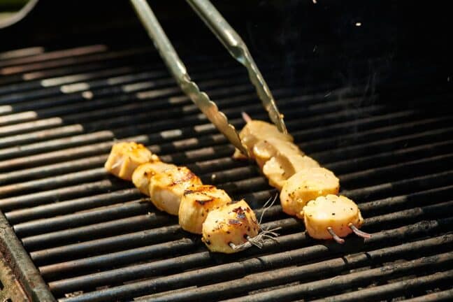 Tongs flipping skewered scallops on a grill.
