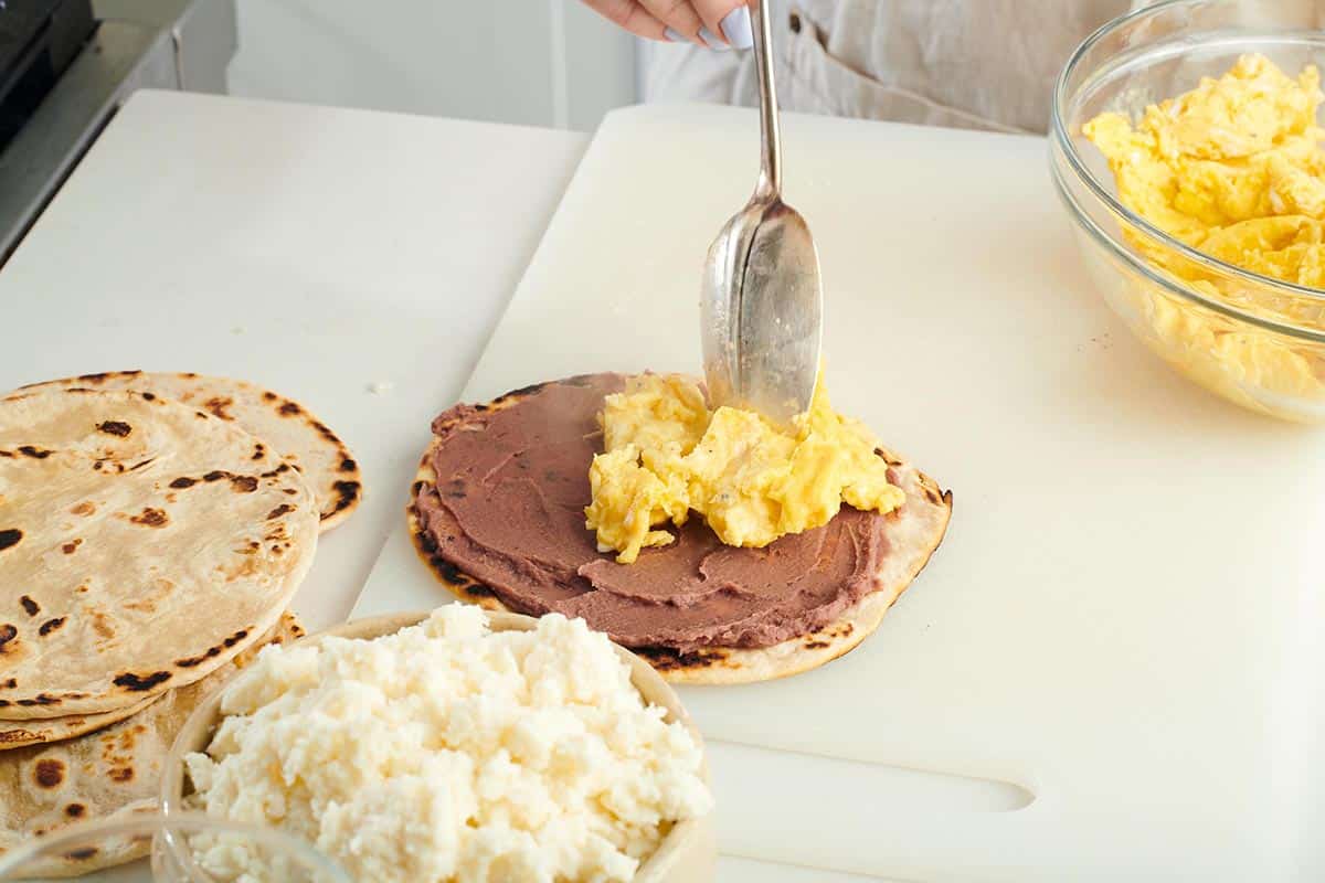Woman spooning scrambled eggs onto a flour tortilla spread with refried beans.