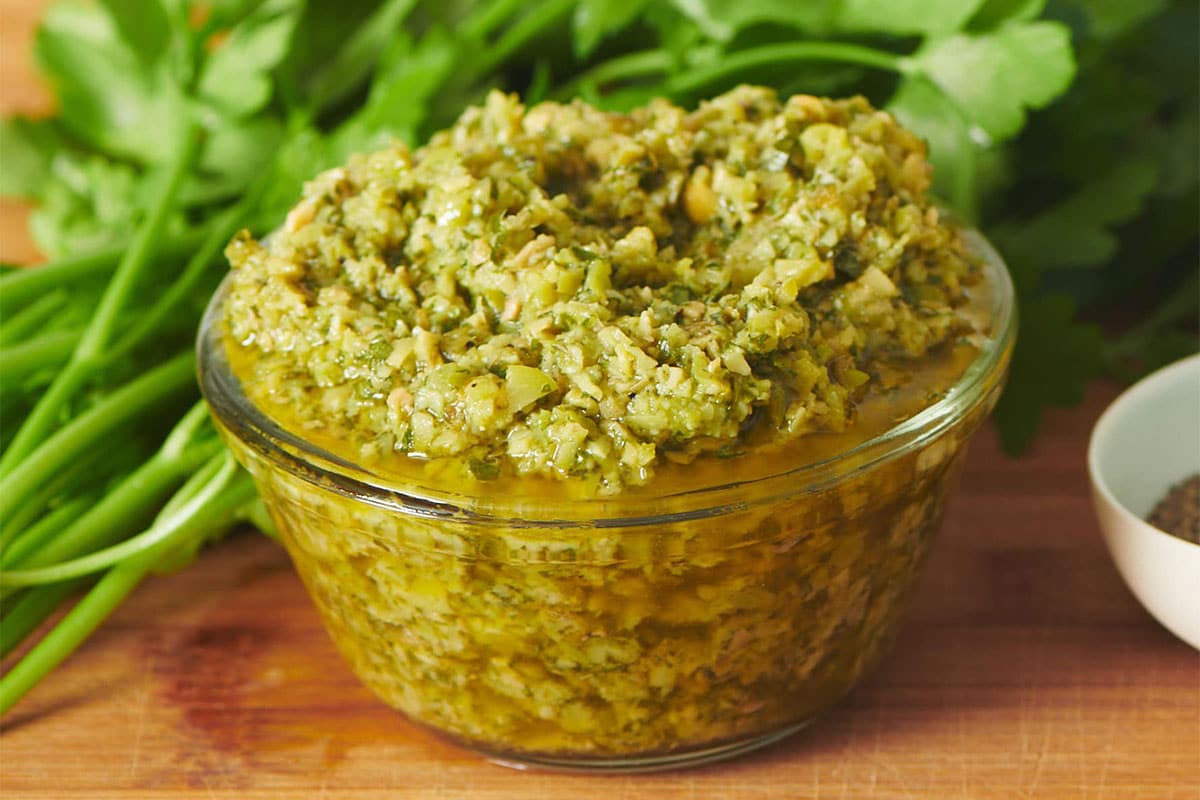 Green olive tapenade in glass bowl on table with herbs.
