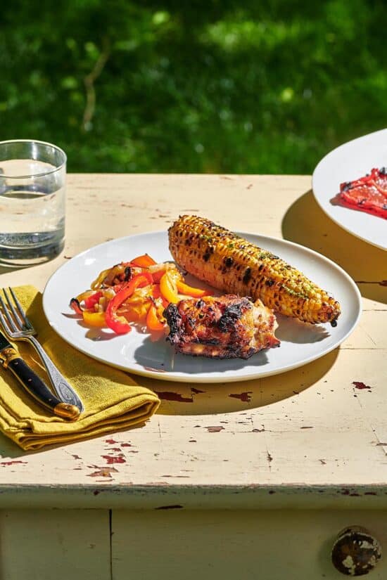 Grilled Chicken Thighs on a plate with corn and peppers set on an outdoor table.