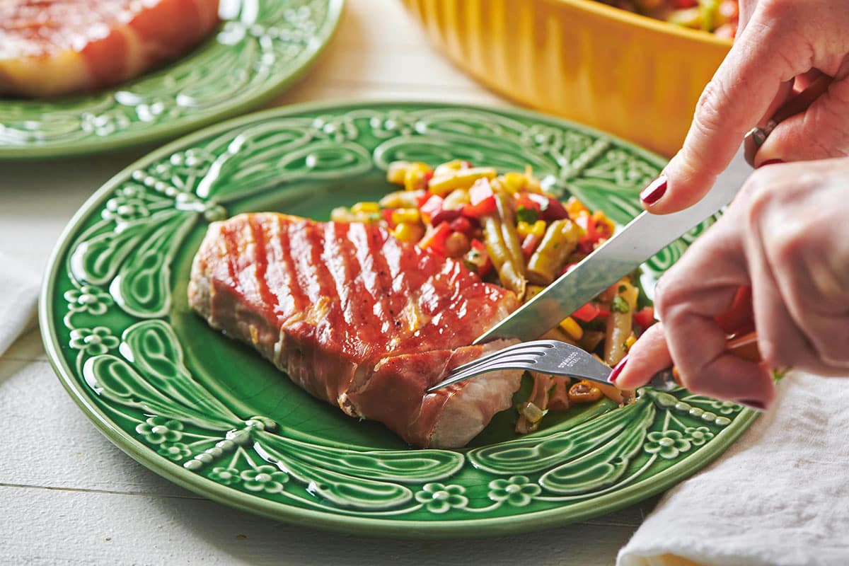 Woman using a fork and knife to slice a Grilled Prosciutto Wrapped Pork Chop.