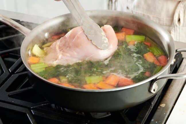 Tongs placing chicken into a pan of water, herbs, and vegetables.