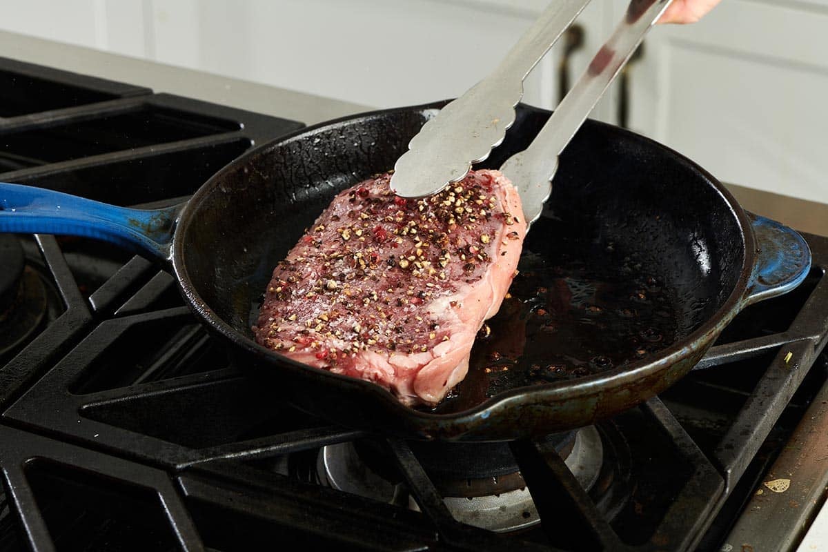Tongs flipping a seasoned steak in a pan.