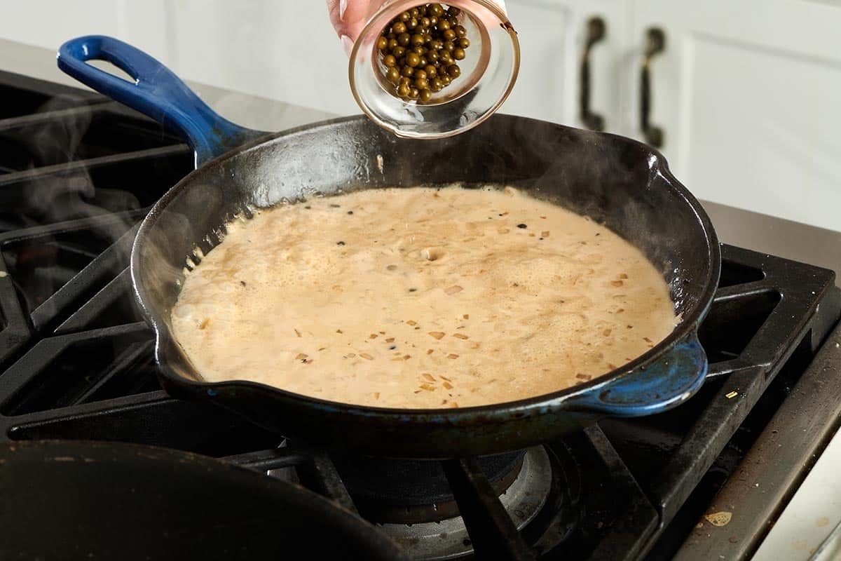 Green peppercorns being poured into a creamy sauce.