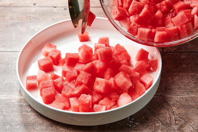 Tongs placing diced watermelon into a bowl.