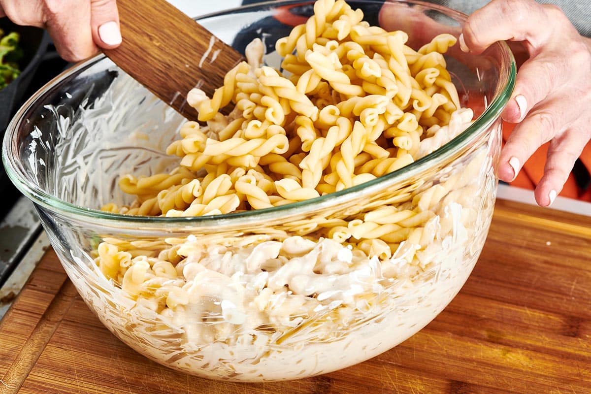 Wooden spatula stirring cooked noodles into a bowl of white sauce.
