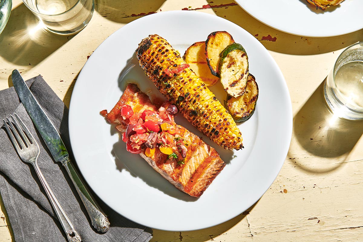 Grilled salmon, corn, and eggplant with water glasses on picnic table.