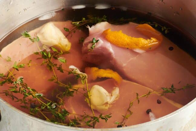Pork chops floating in a pot of brine with thyme, orange peel, and garlic.