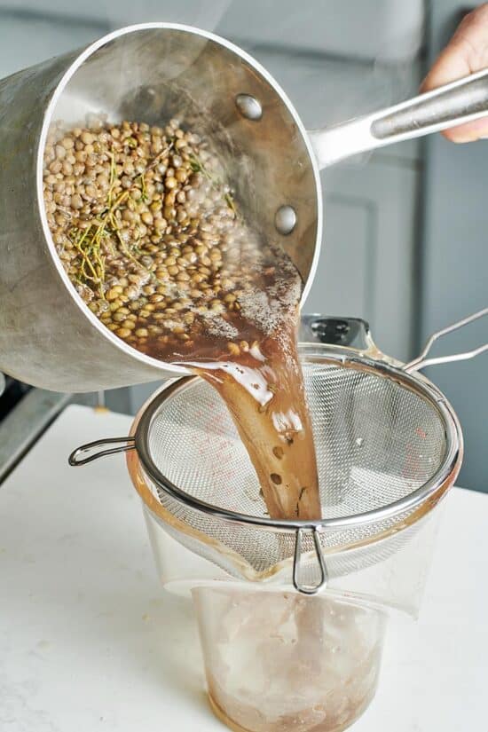 Pot of water, French lentils, and herbs being poured into a sieve.