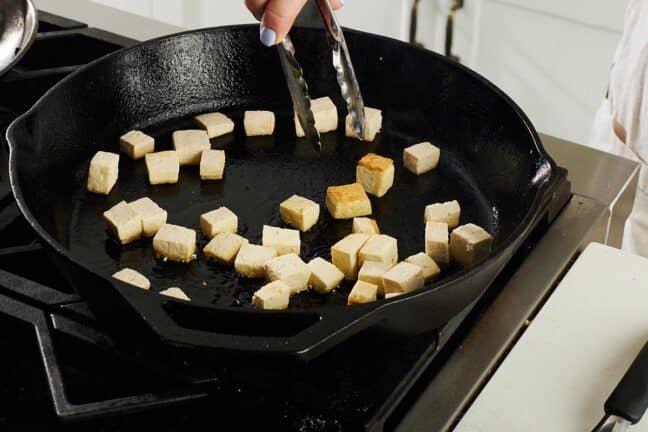 Tongs grabbing cubes of tofu in a cast iron pan.