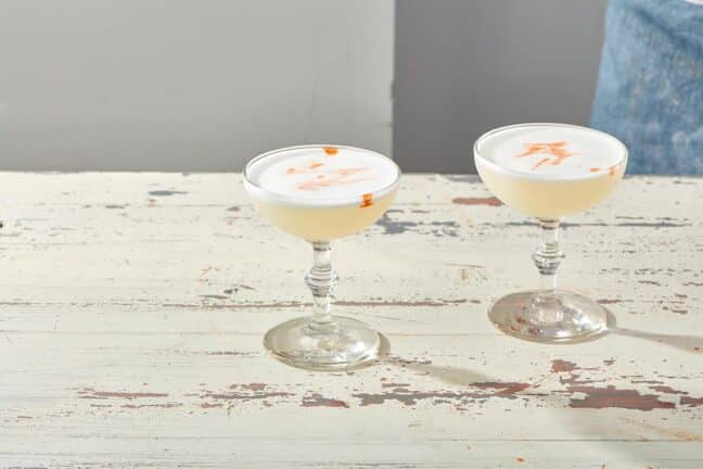 Two Pisco Sours in long-stemmed glasses on a white, wooden table.