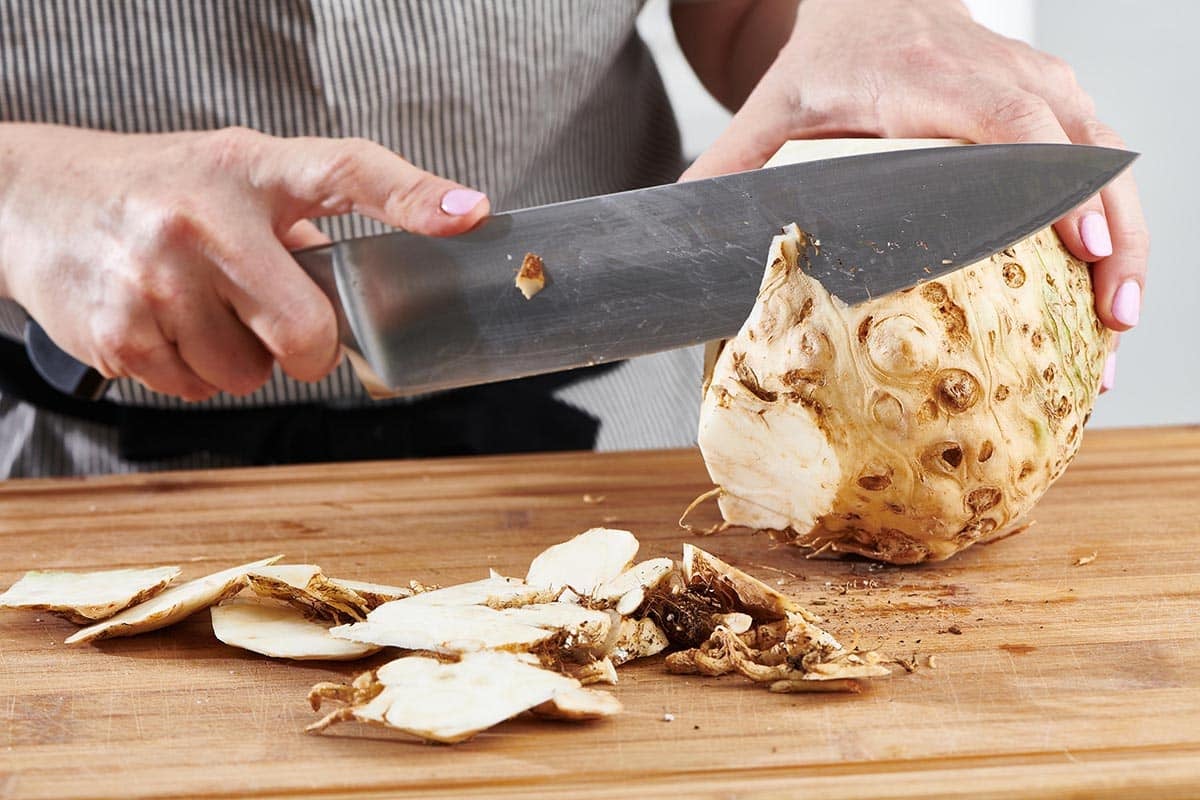 Woman using a large knife to cut the skin off of a celeriac.
