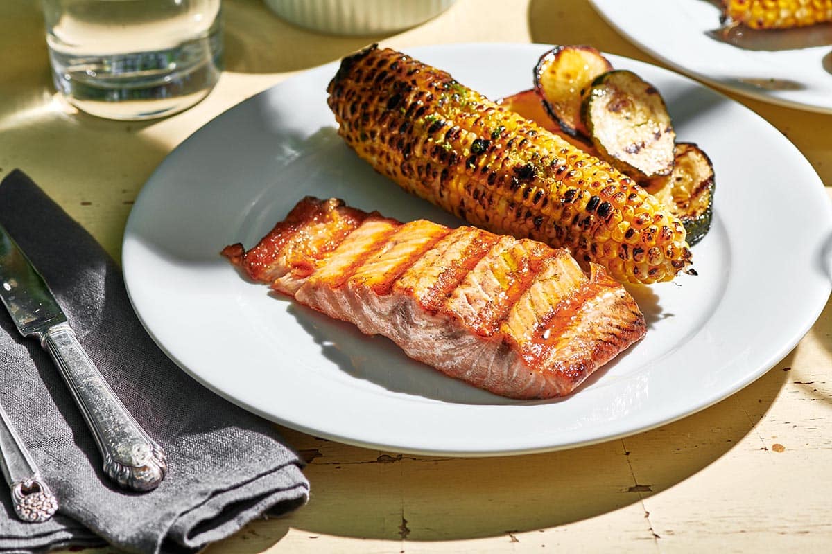 White plate with grilled salmon, corn on the cob, and eggplant on picnic table.
