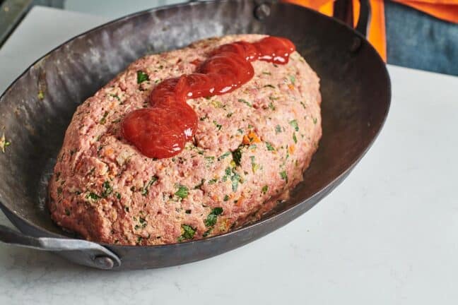 Uncooked Meatloaf in a pan with a drizzle of ketchup on top.