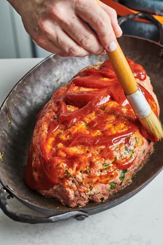 Woman brushing ketchup onto meatloaf in a pan.