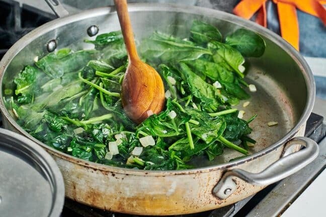 Wooden spoon stirring a skillet of spinach and onions.