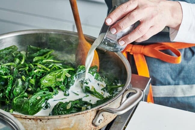 Woman pouring half and half into a skillet of spinach and onions.