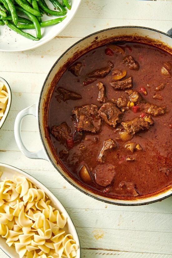 Pot of Hungarian Goulash next to dishes of egg noodles.