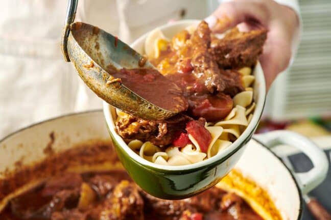 Large ladle serving Hungarian Goulash into a dish of egg noodles.