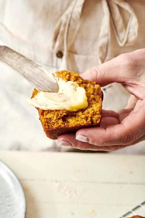 Woman using a butter knife to spread butter onto a mini pumpkin bread loaf.