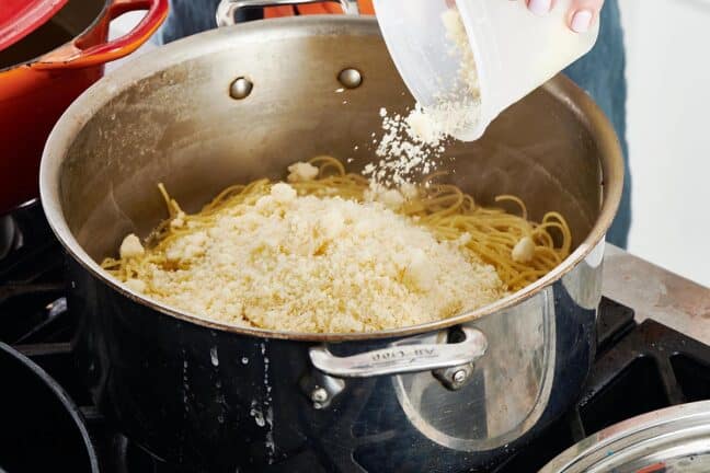 Container of parmesan cheese pouring into a pot of noodles.