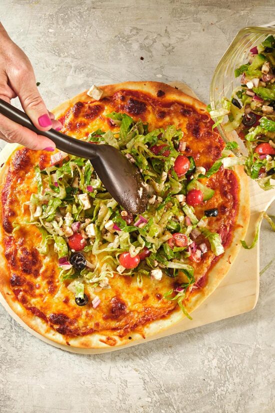 Woman using a spoon to scoop Greek Salad onto a Greek Salad Pizza.