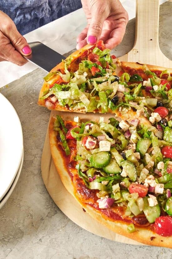 Woman using a spatula to serve a slice of Greek Salad Pizza.