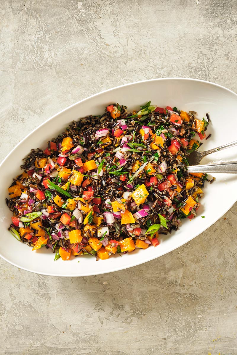 Two spoons in a oblong bowl of colorful Wild Rice and Sweet Potato Salad.