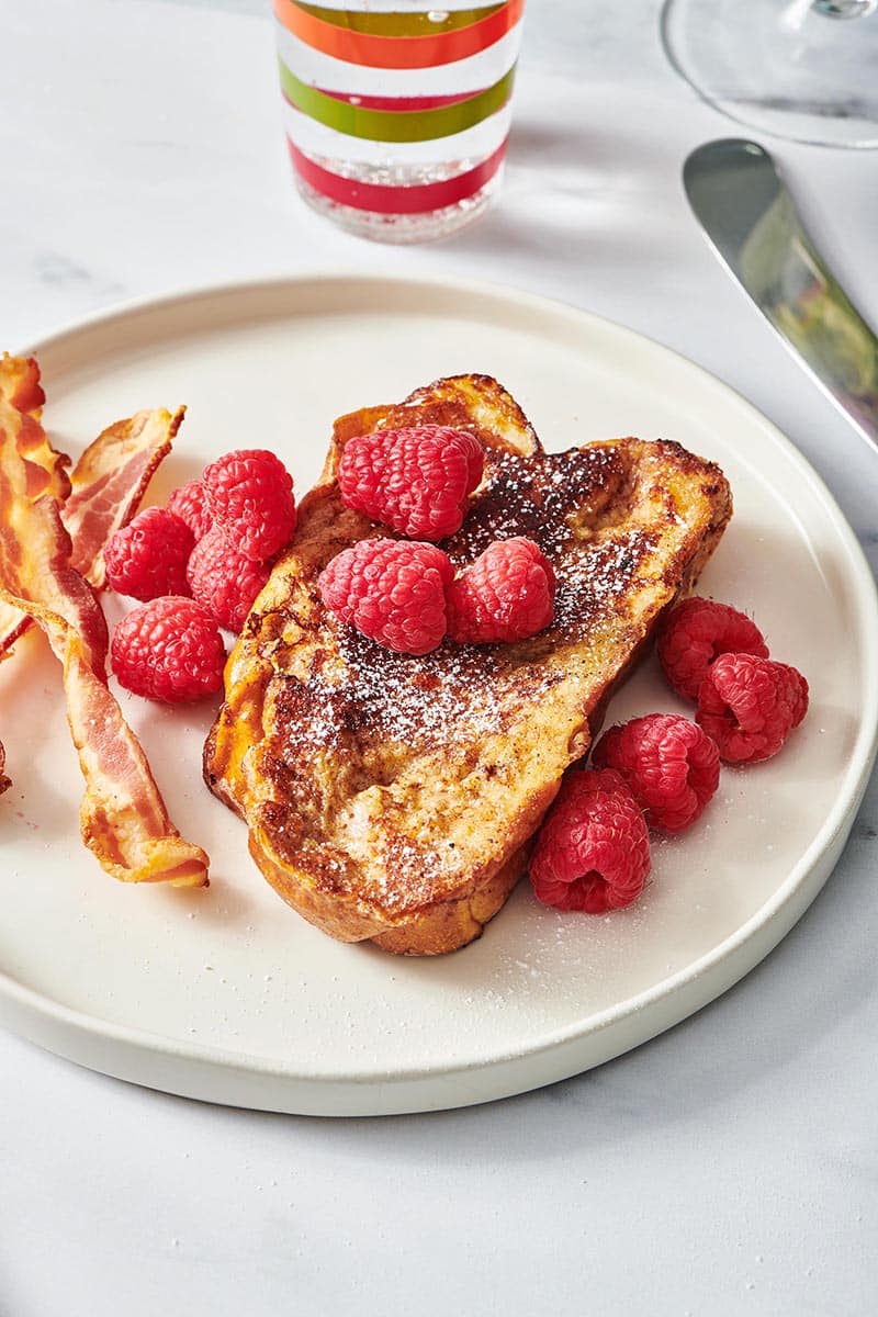 Challah French Toast topped with raspberries with bacon on plate.