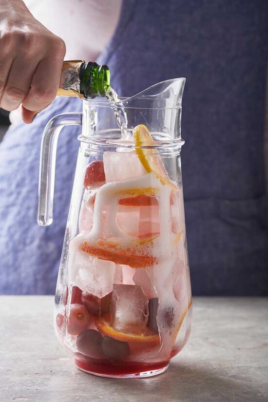 Sparkling wine pouring into a pitcher of ice and fruit.