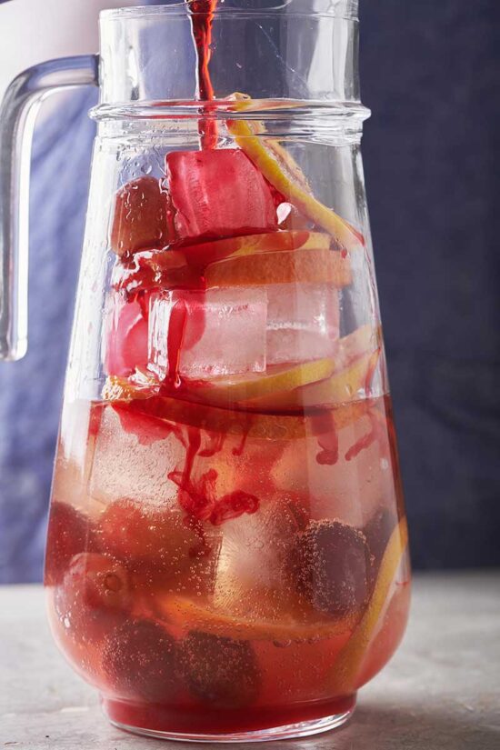 Red hibiscus tea being poured into a pitcher of ice, fruit, and other ingredients.