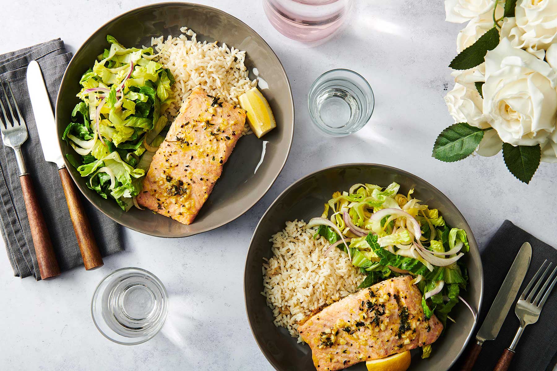 Two dark plates of baked salmon, salad, and rice on a white table.