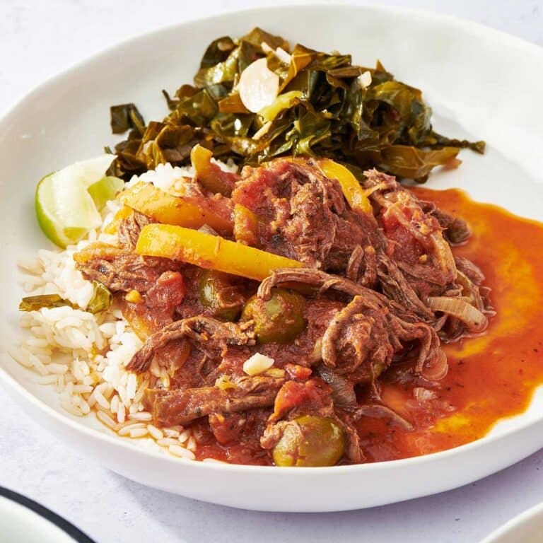 White plate with Ropa Vieja over rice next to cooked greens.