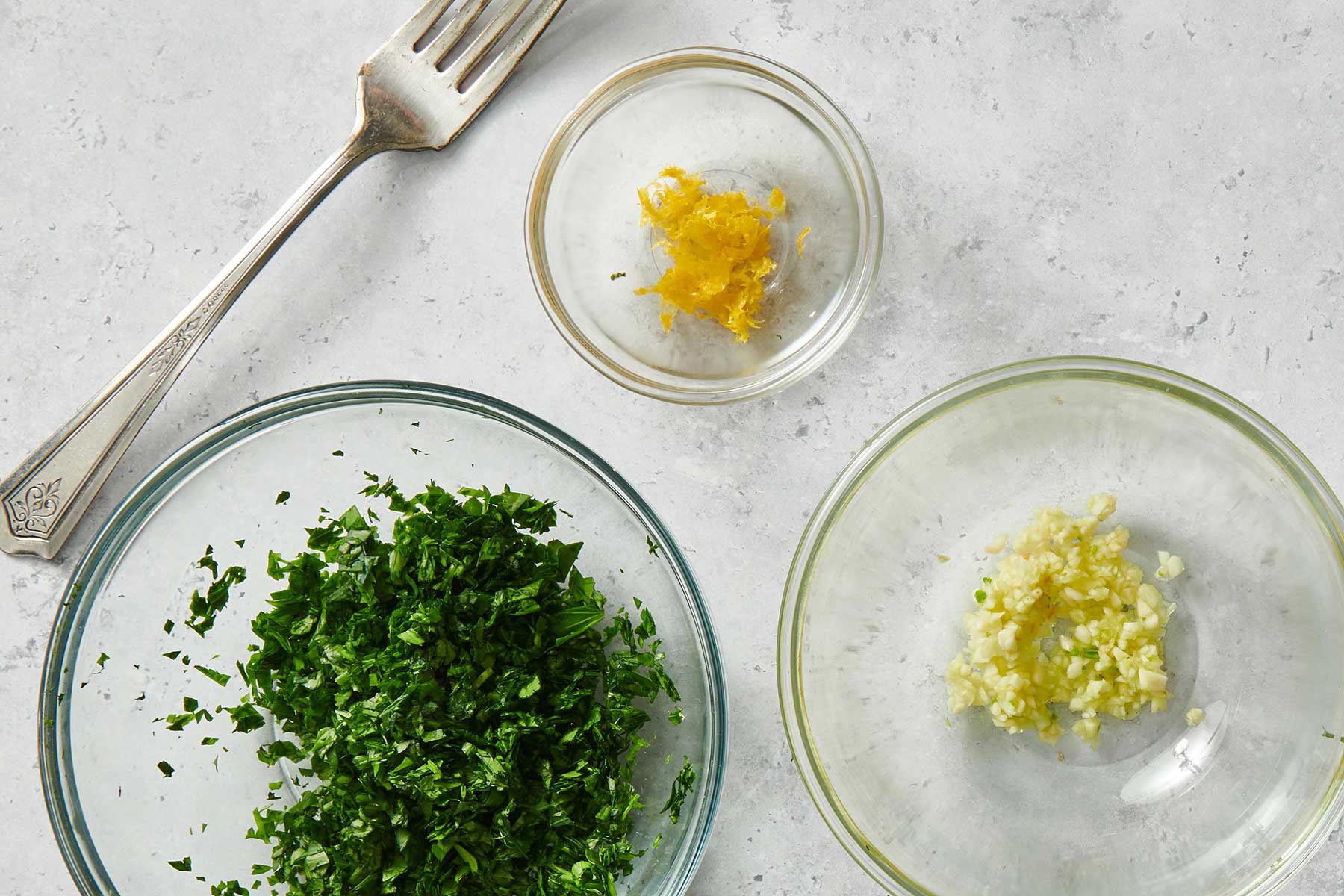 Glass bowls of parsley, lemon zest, and chopped garlic.